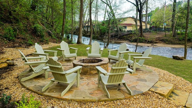Ein runder Sitzbereich mit Adirondack-Stühlen um ein Steinkaminfeuer, auf einer gepflasterten Terrasse mit Kies, mit Blick auf einen Fluss und umgeben von Bäumen.