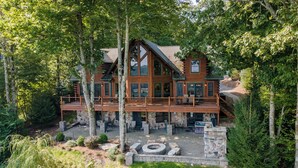 A large log cabin with a wooden deck, stone fire pit, and surrounded by dense forest. - Designer Mountain Lodge w/ Panoramic Views & Sauna (McHenry)