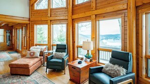 A spacious room with large windows, wooden walls, a sofa, two armchairs, a small table with a lamp, and a decorative rug. - Designer Mountain Lodge w/ Panoramic Views & Sauna (McHenry)