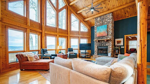 A spacious living room with large windows, a stone fireplace, and wooden beams. - Designer Mountain Lodge w/ Panoramic Views & Sauna (McHenry)