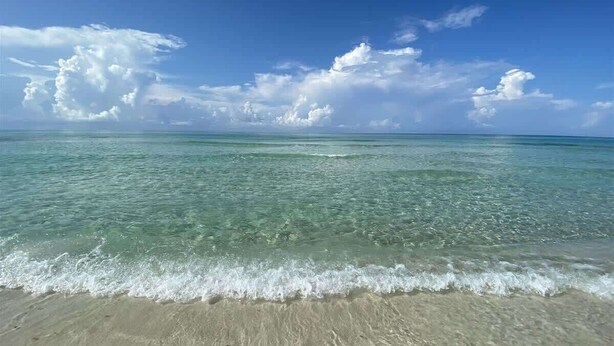 A beach with clear water, sandy shore, and a blue sky with clouds.