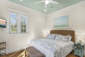Quarto com cama, mesa de cabeceira, banquinho, ventilador de teto e um quadro na parede.