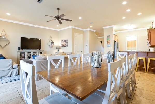 A spacious living area with a wooden dining table, white chairs, a ceiling fan, a flat-screen TV, and a kitchen in the background.