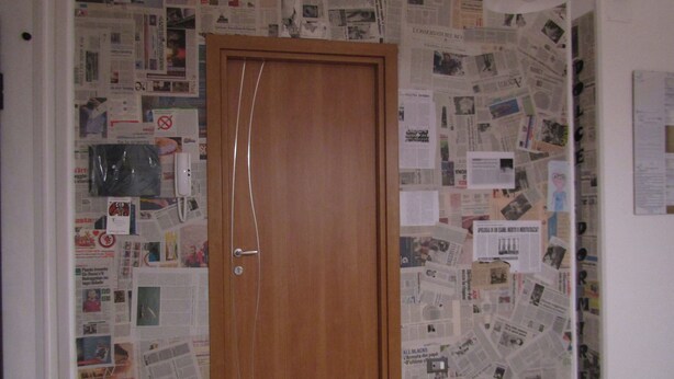 A room with a wooden door, walls covered with various papers and photographs, and a ceiling light fixture. - B&B Dolce Dormire (Foggia)