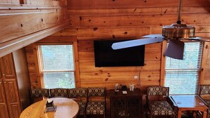A wooden interior with a round dining table, patterned chairs, a flat-screen TV mounted on the wall, and a ceiling fan. - 'Just a Step Away' - a Secluded Cabin on Stover Creek w/Fiber Wi-Fi & Hot Tub (Ellijay)