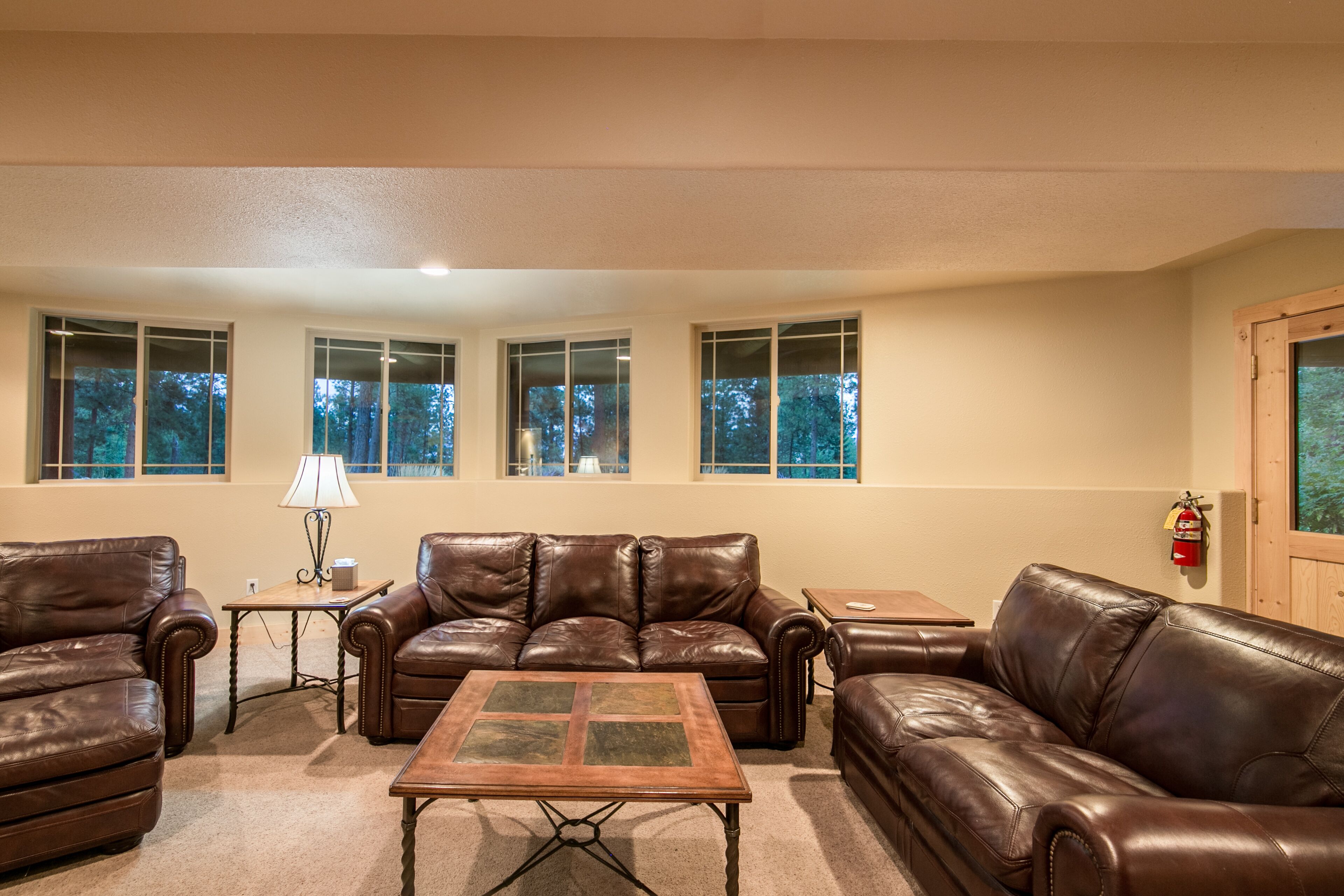 A living room with leather sofas, a wooden coffee table, and a lamp. There are windows with views of trees.
