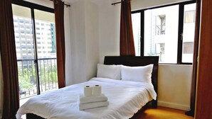 A neatly made bed with white linens and two rolls of toilet paper, a window with brown curtains, and a view of a building outside. - Forbeswood Heights 2BR by Stays PH (Taguig)