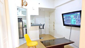 A modern living room with a TV, a yellow chair, a wooden coffee table, and a kitchen area visible through an open door. - Forbeswood Heights 2BR by Stays PH (Taguig)