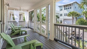 A covered porch with green chairs and a table, a ceiling fan, and a view of a house and palm trees.