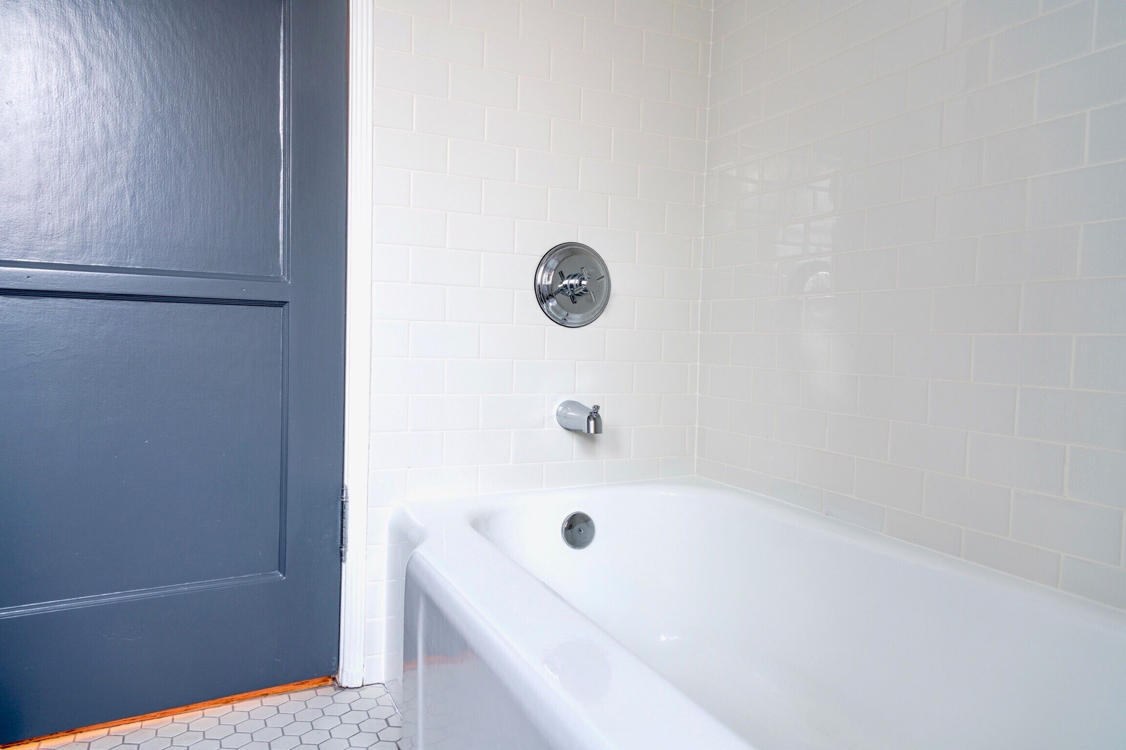 A bathroom with a white bathtub, a gray door, and white tiled walls.