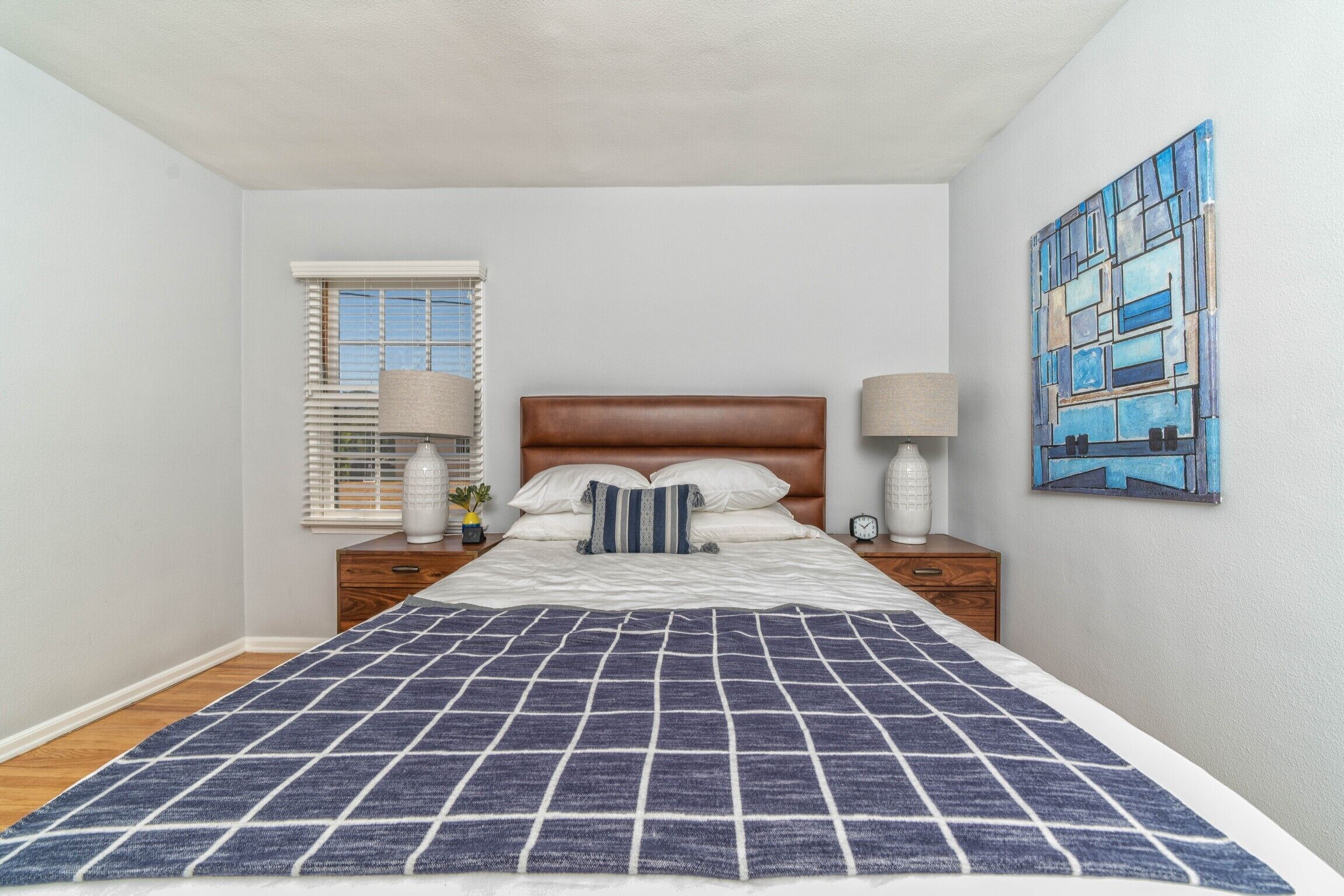 Une chambre à coucher avec un lit, des tables de chevet, une fenêtre avec des stores et un tableau au mur.