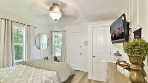 Ein Schlafzimmer mit einem Bett, einem Deckenventilator, einem an der Wand befestigten Fernseher und einem Fenster mit Vorhängen.