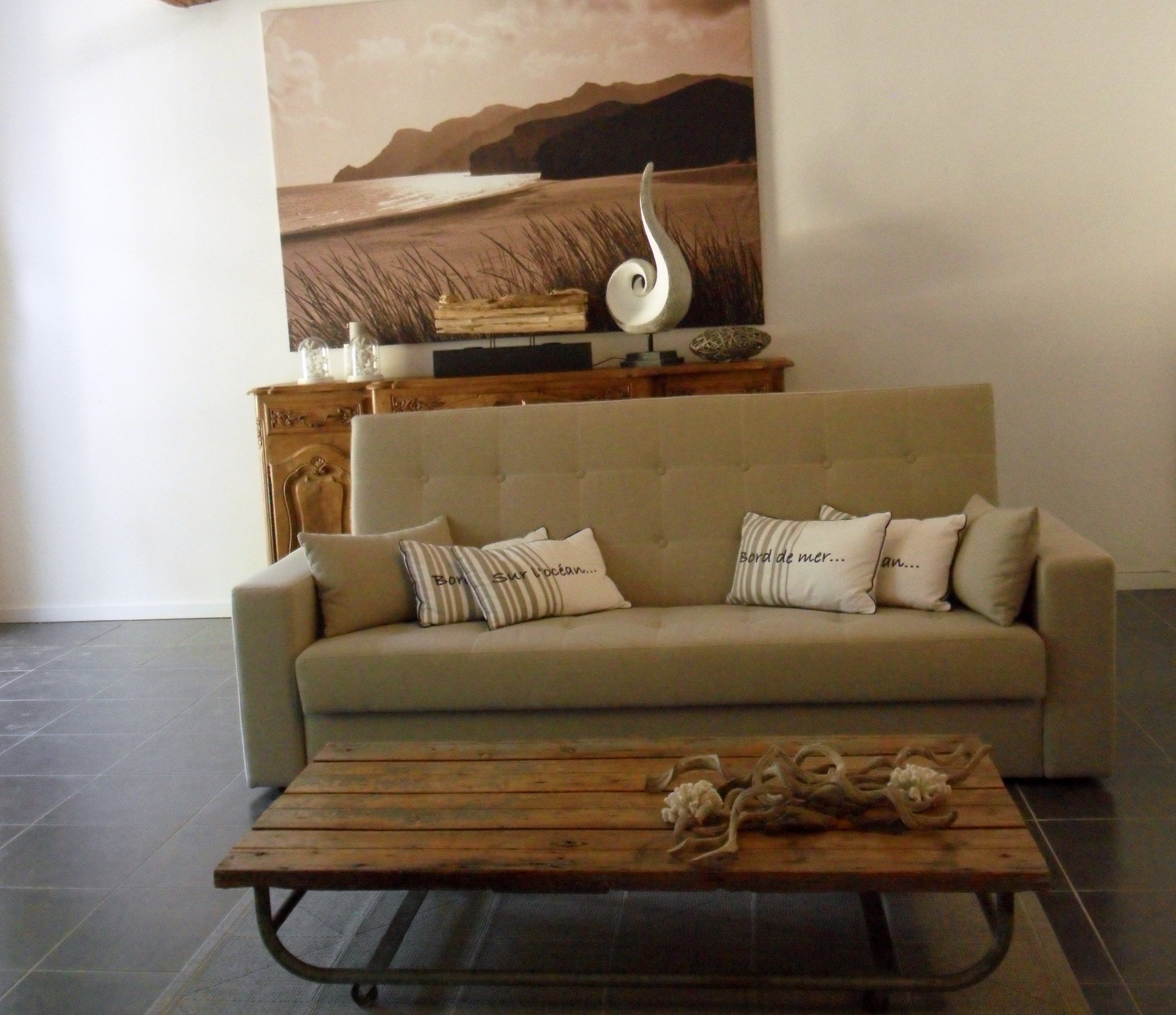 Una sala de estar con un sofá color beige, almohadas a rayas, una mesa de centro de madera y un cuadro grande de un paisaje marino.