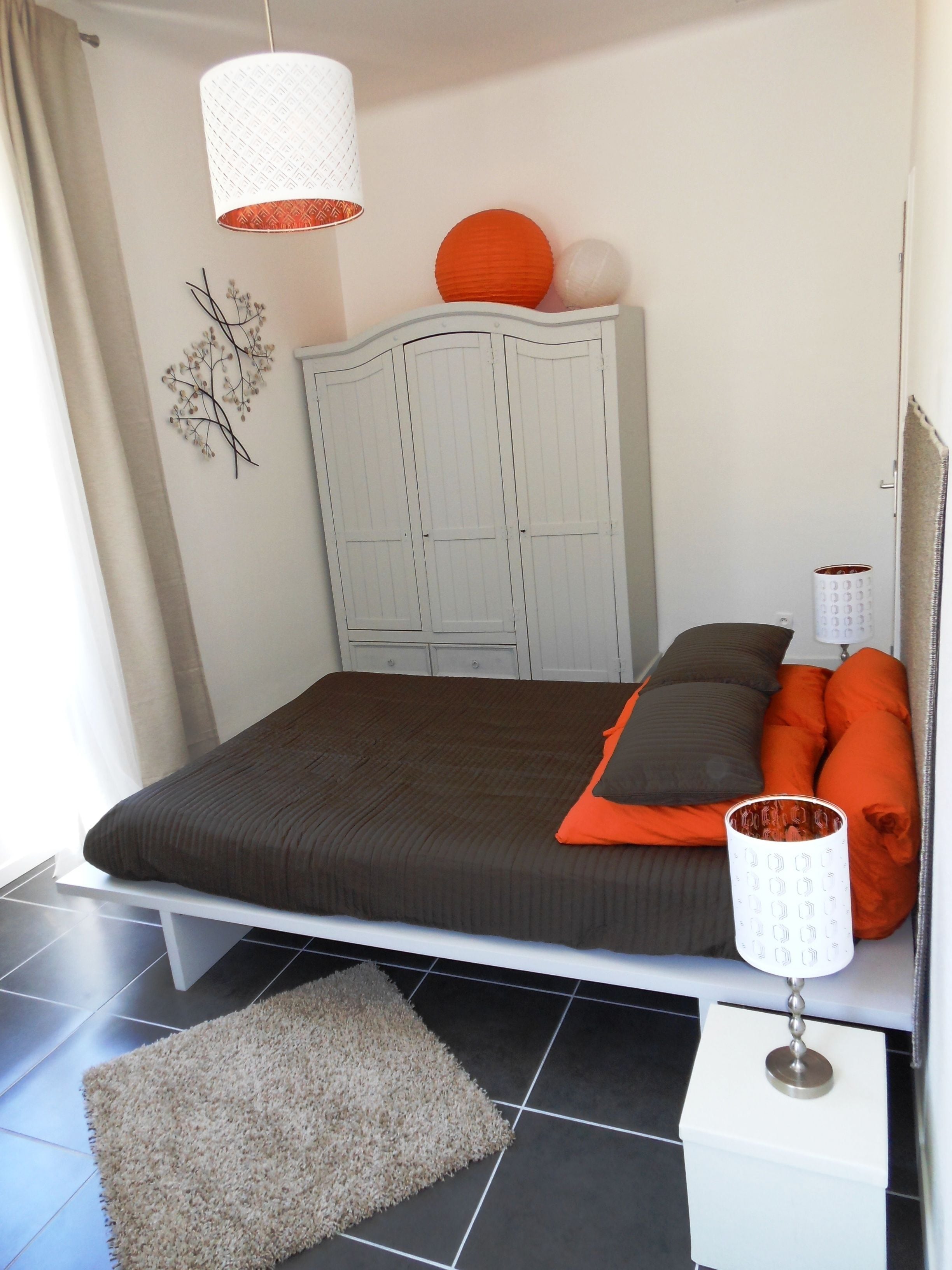 A bedroom with a bed, a nightstand, a lamp, a wardrobe, and a decorative wall piece.
