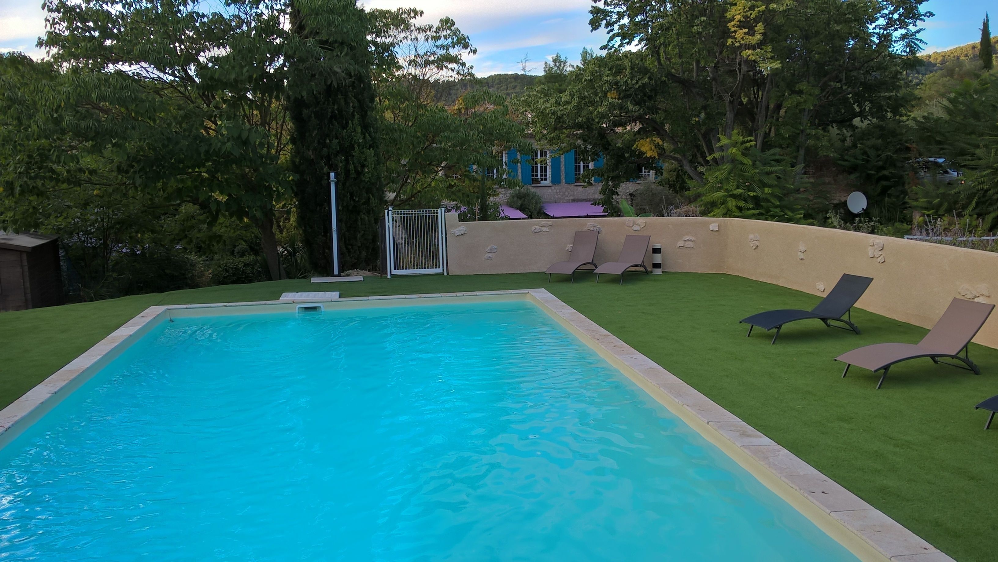Una piscina rettangolare con superficie d'acqua blu trasparente, circondata da erba sintetica verde. Si notano diversi lettini da relax e un'area salotto coperta con tetto viola.
