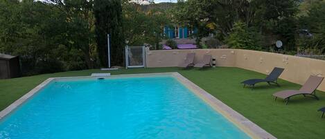 Una piscina rettangolare con superficie d'acqua blu trasparente, circondata da erba sintetica verde. Si notano diversi lettini da relax e un'area salotto coperta con tetto viola.