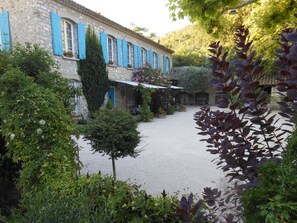 Un bâtiment en pierre avec des volets bleus, entouré de verdure et doté d’une cour recouverte de gravier.
