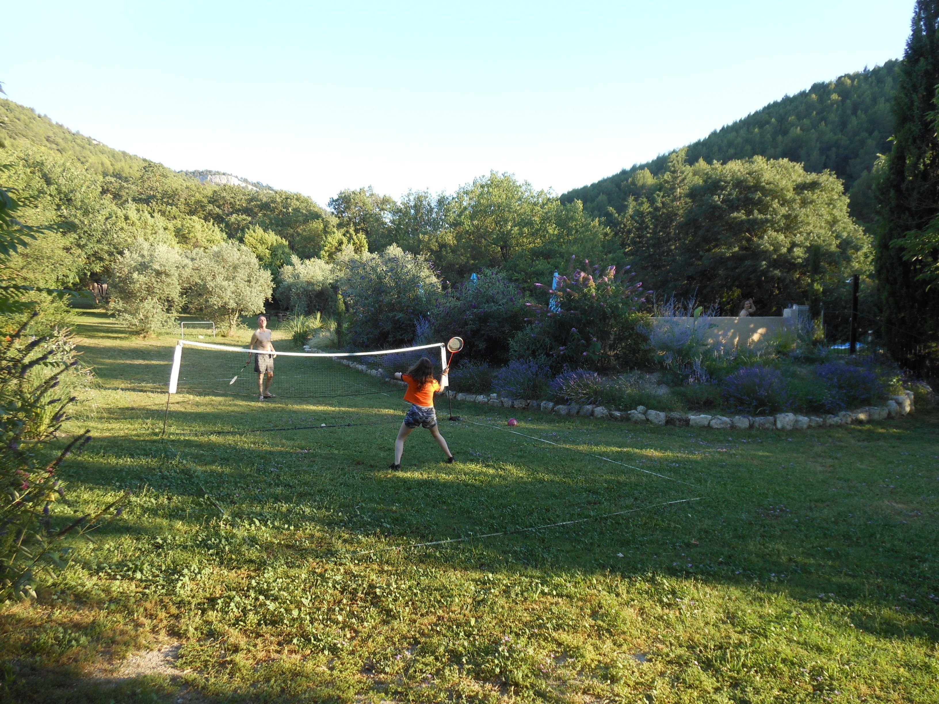 A grassy outdoor area with a netted tennis court, surrounded by trees and hills.