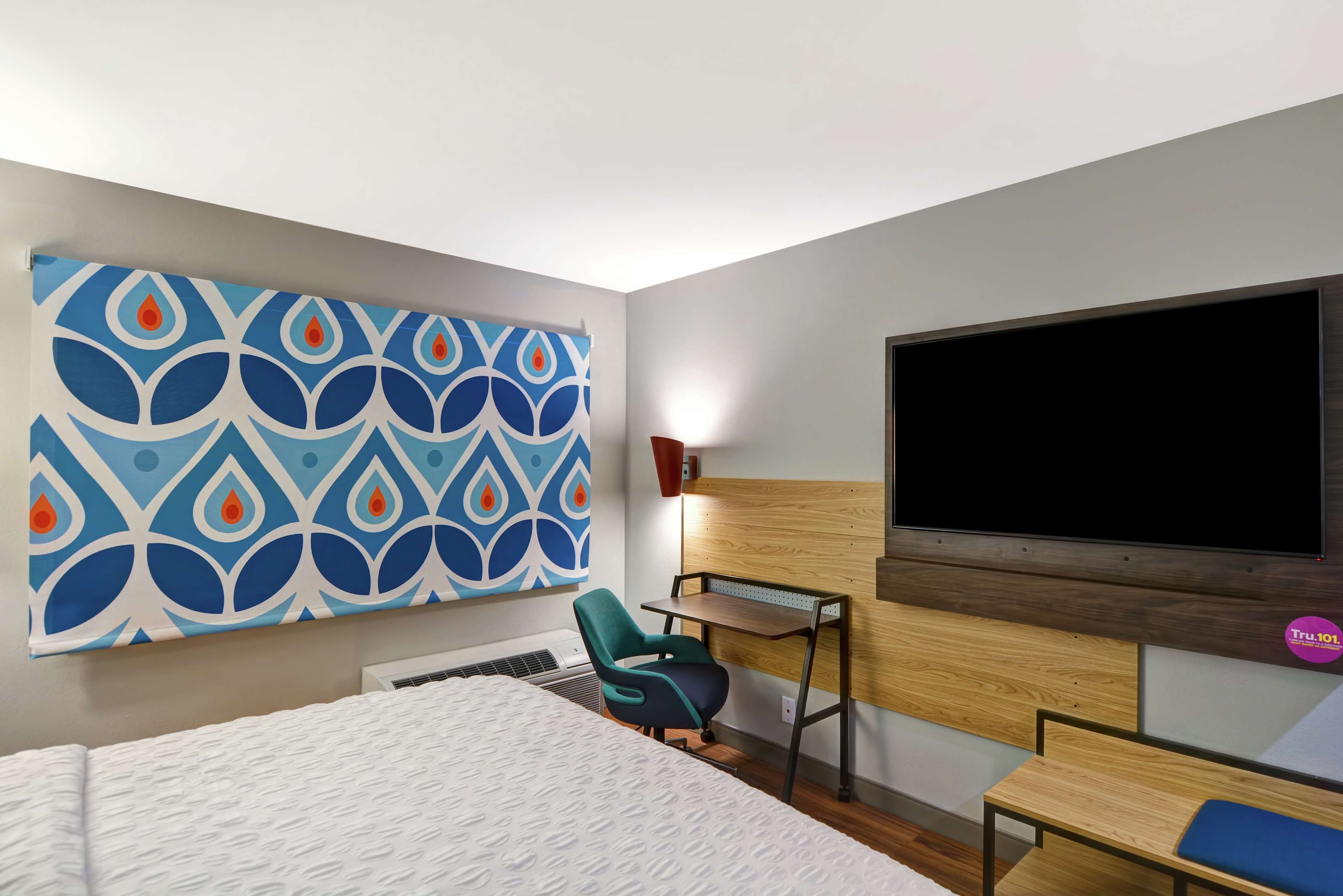 A hotel room with a bed, a desk, a chair, a TV, and a wall art piece.