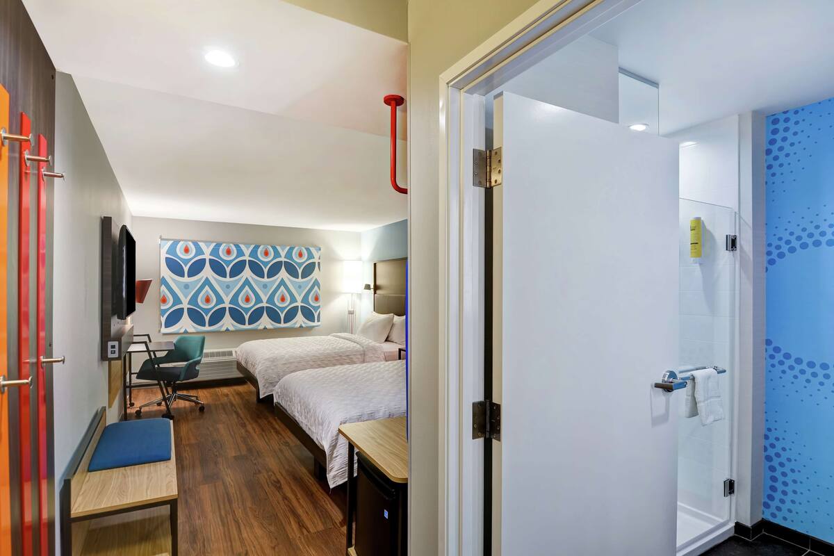 A hotel room with two beds, a desk, a chair, a TV, and a bathroom with a shower and toilet.