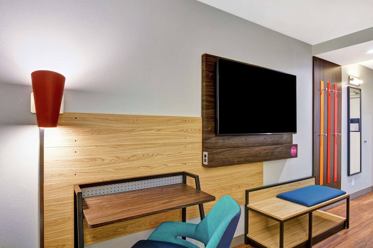 A modern hotel room with a wooden wall panel, a flat-screen TV, a small table, a blue cushioned chair, and a red floor lamp.