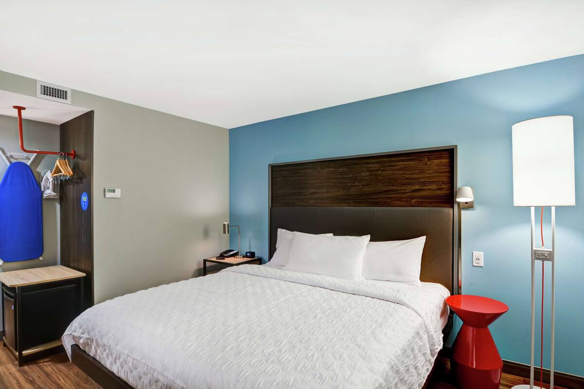 A hotel room with a large bed, a bedside table, a nightstand, a red accent chair, a floor lamp, and a wall-mounted heater.