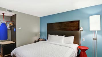 A hotel room with a large bed, a bedside table, a nightstand, a red accent chair, a floor lamp, and a wall-mounted heater.