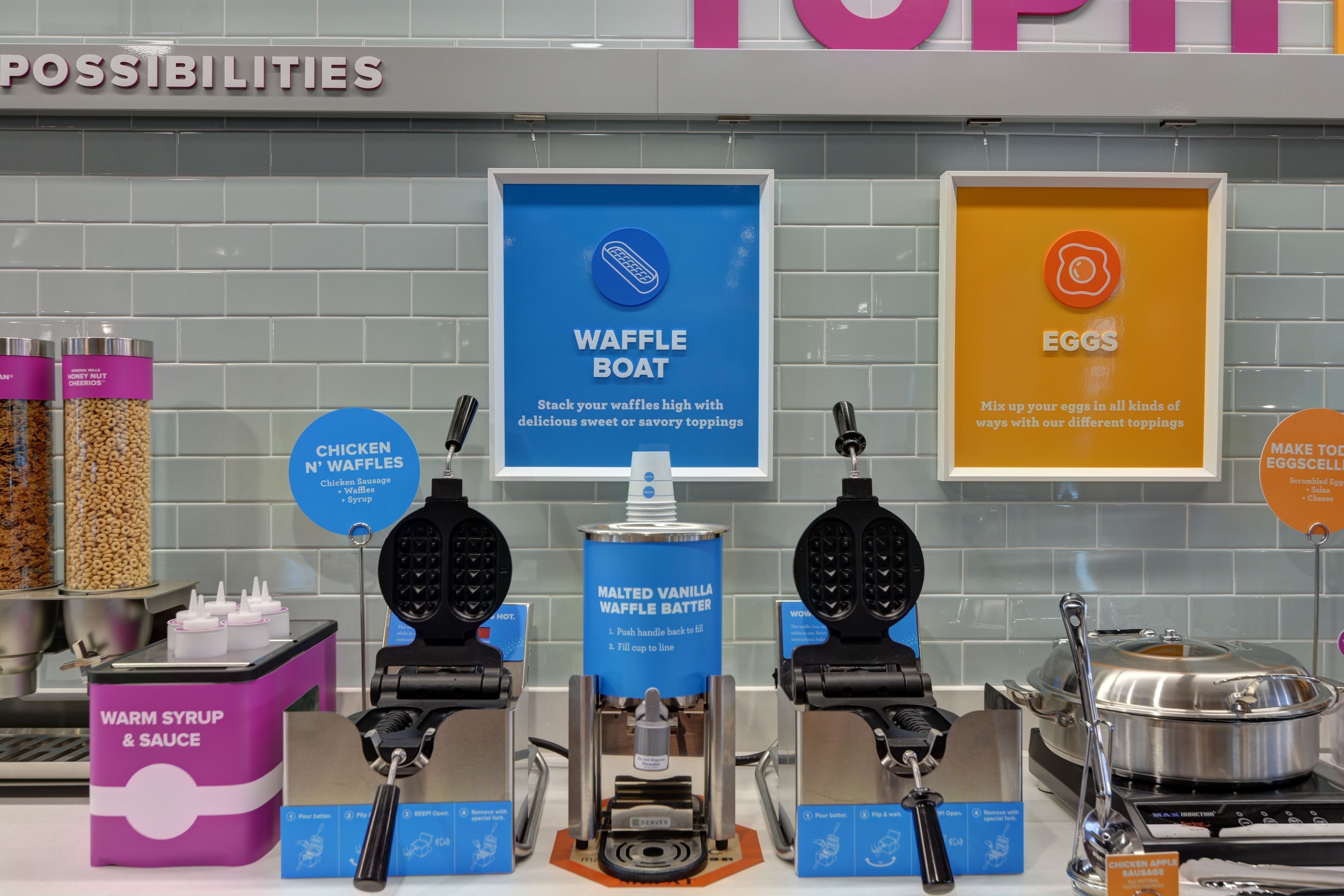 A waffle station with waffle boats, a malted vanilla waffle batter dispenser, and a sign for chicken sausage waffles.