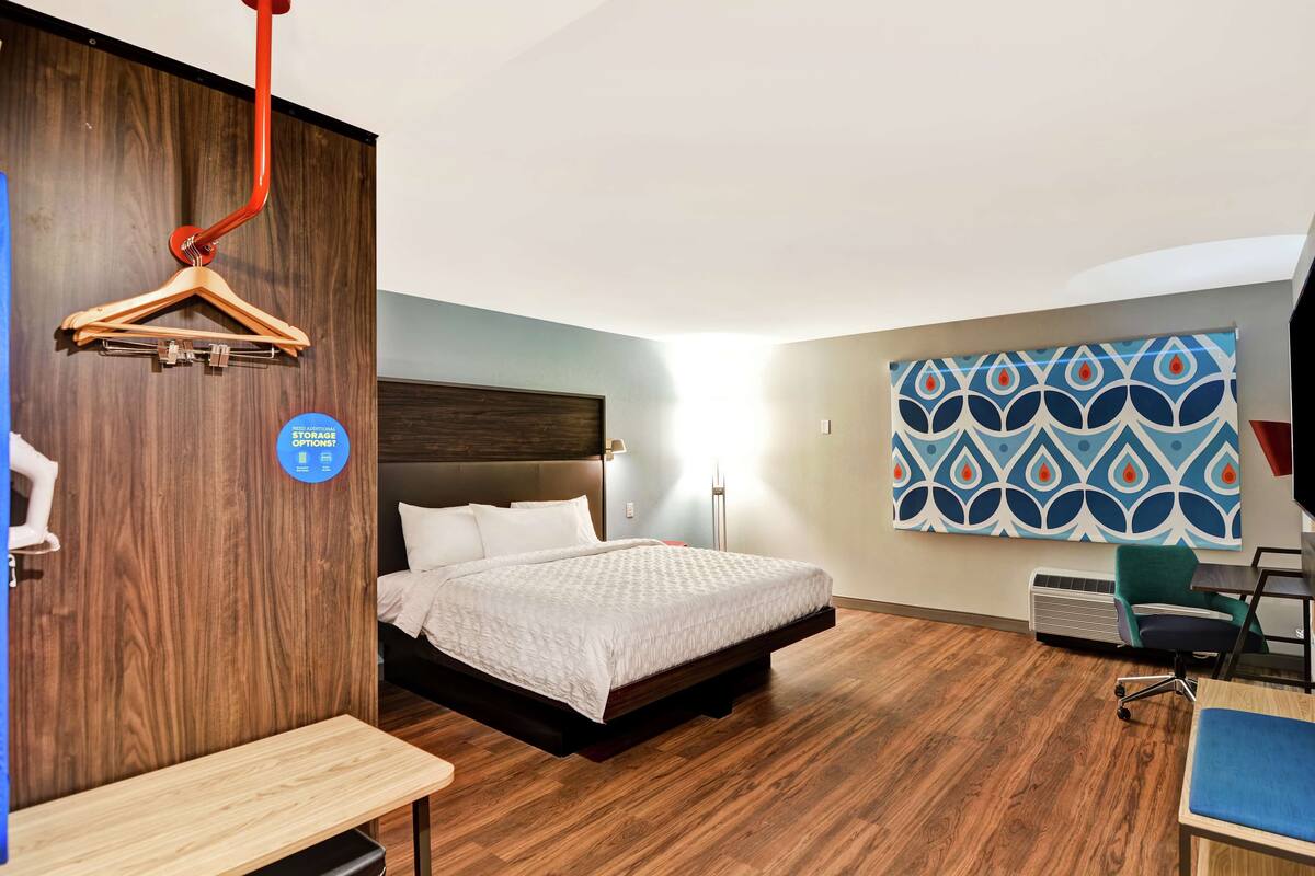 A hotel room with a bed, a desk, a chair, a hanging coat rack, and a wall-mounted air conditioner.