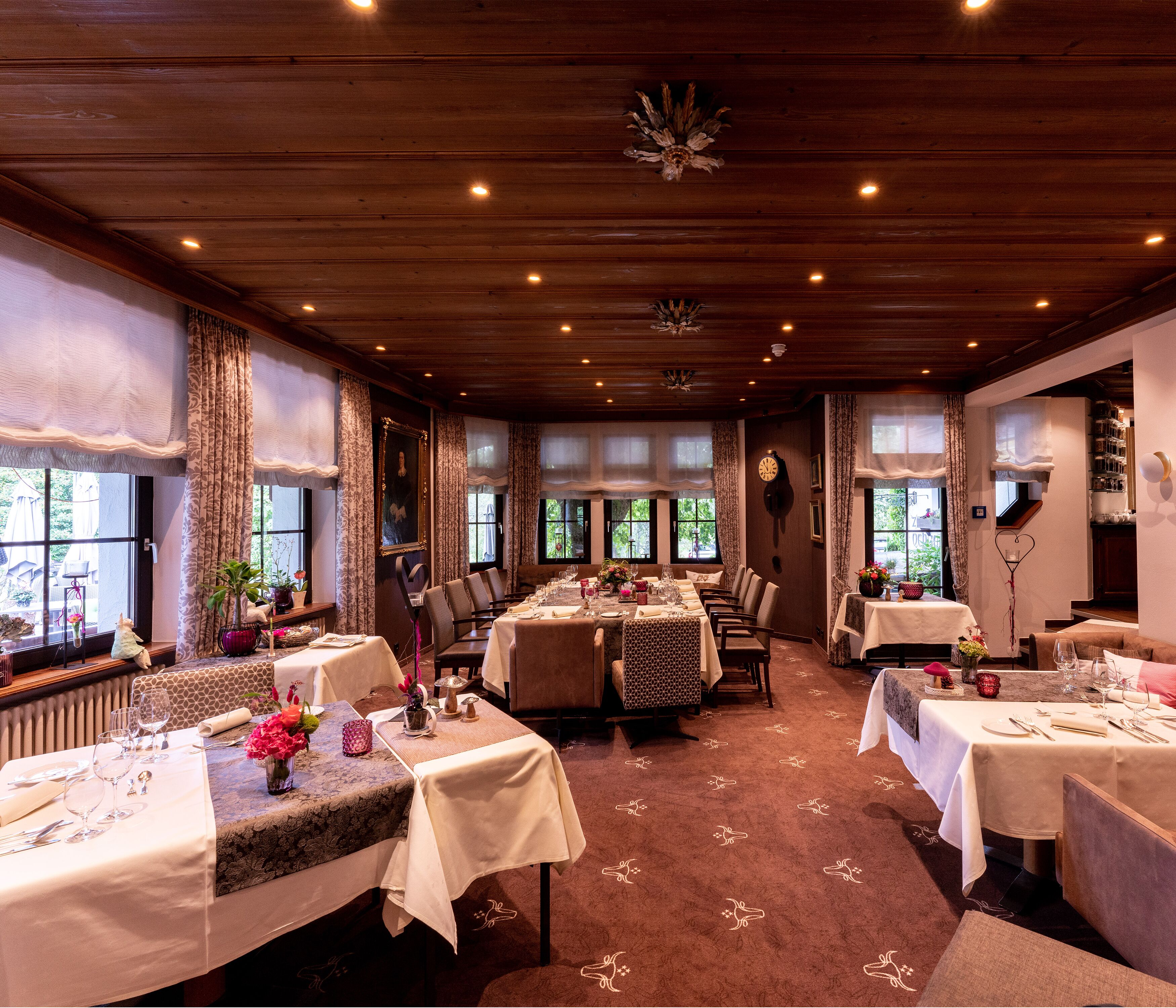 A restaurant with multiple tables set for dining, wooden ceiling, and large windows allowing natural light.