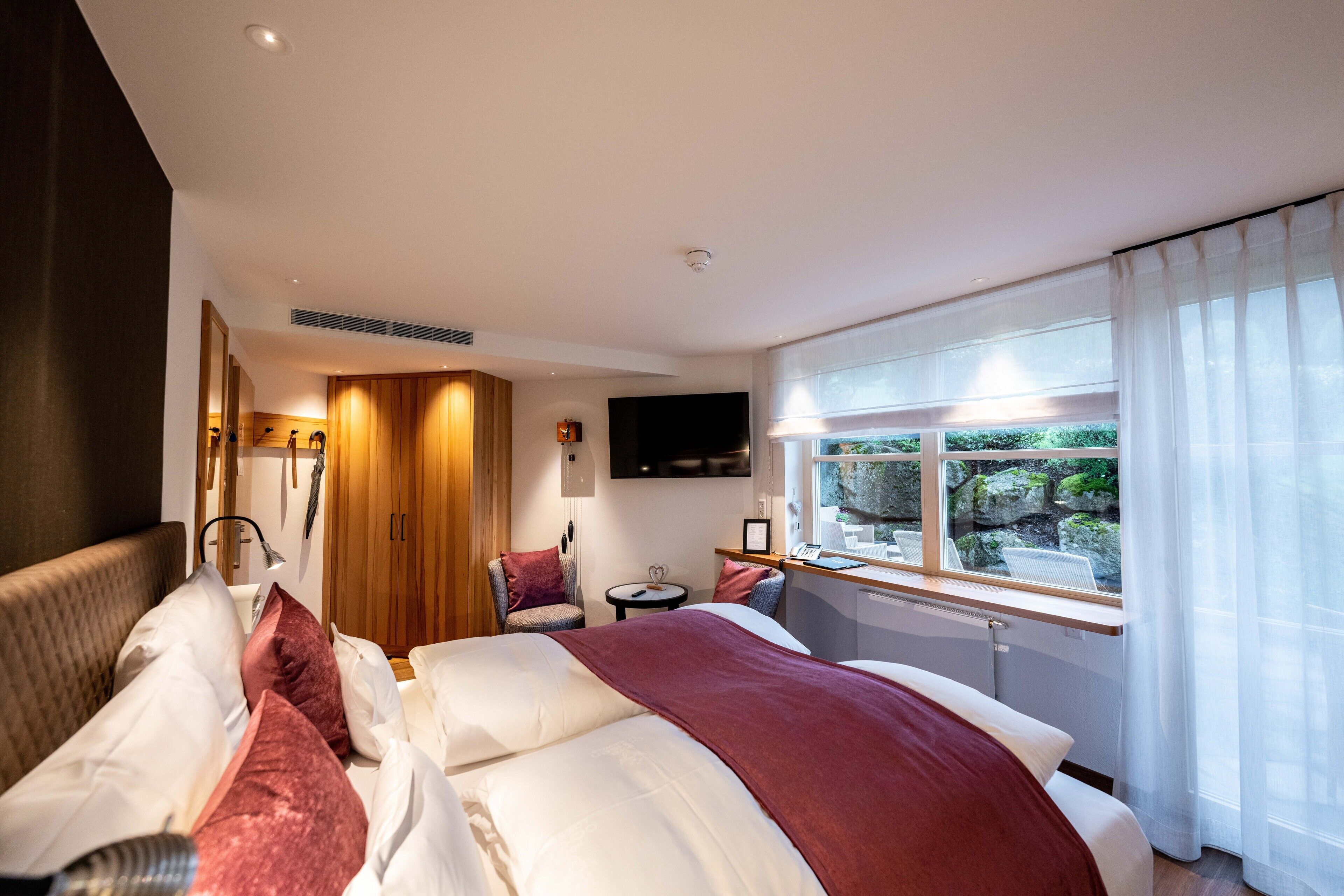 A modern hotel room with a large bed, a sitting area with a chair and a small table, a TV mounted on the wall, and a window with a view of greenery.
