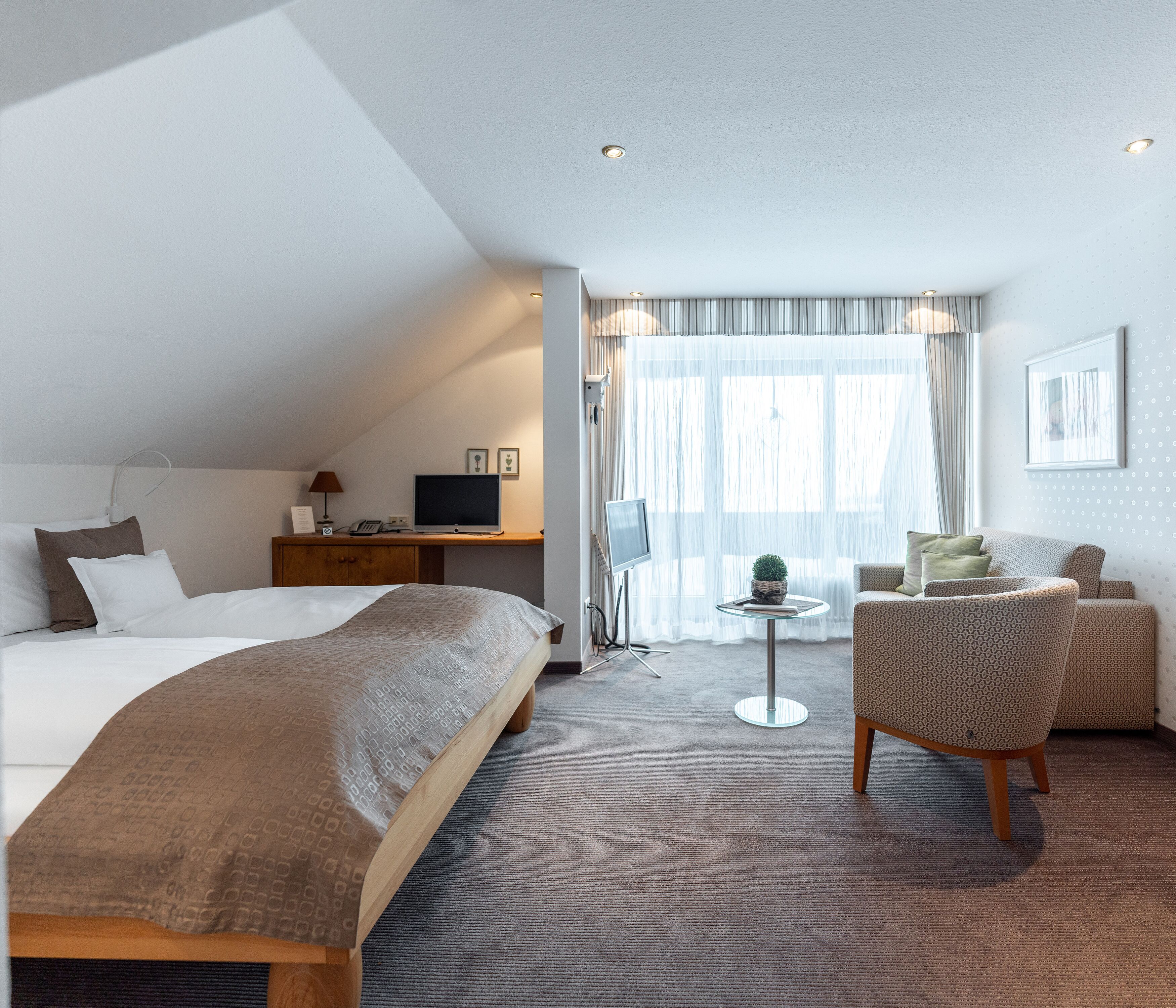A hotel room with a large bed, a desk with a computer, a small table with a plant, and a seating area with chairs.