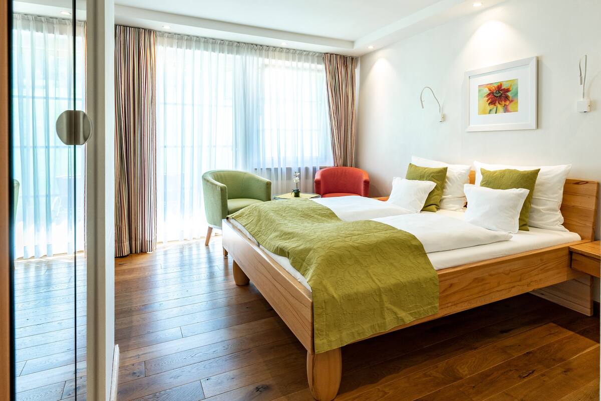 A modern hotel room with a wooden bed, green and white bedding, a red chair, and a large window with sheer curtains.