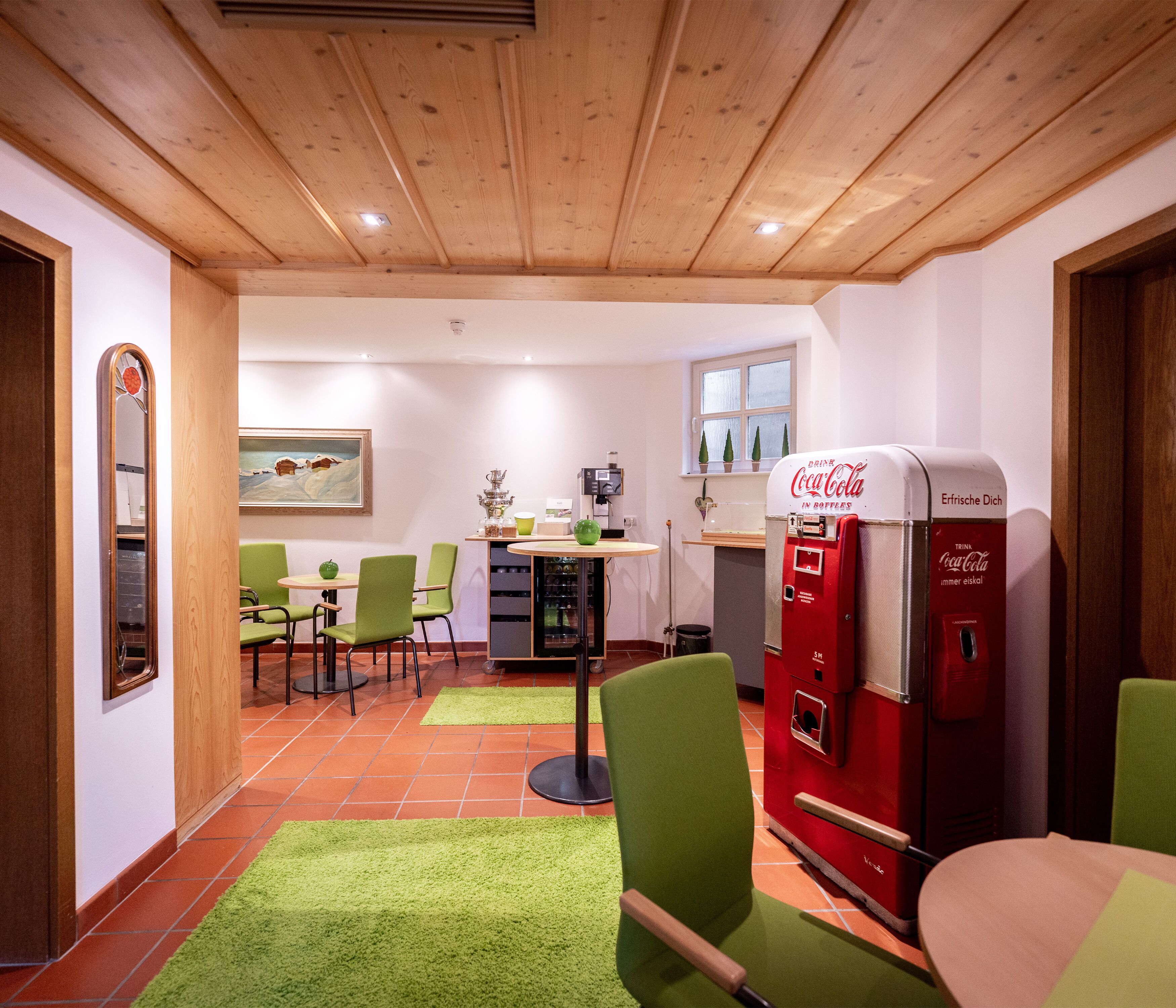 A dining area with green chairs, a red Coca-Cola vending machine, and a painting on the wall.