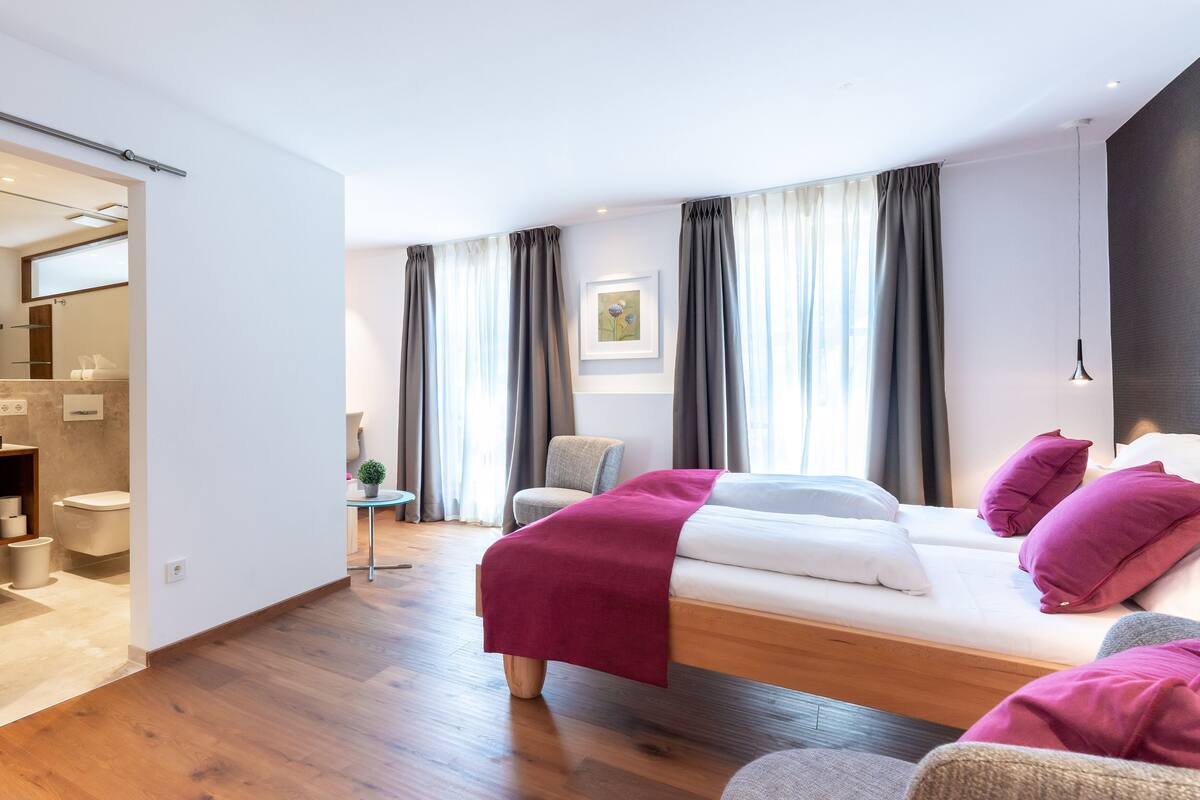 A modern bedroom with a bed, a chair, a small table, and a bathroom visible through an open door.