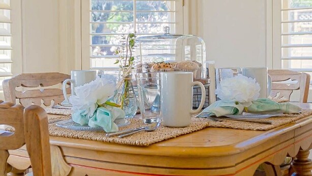 Una mesa de comedor de madera con una cafetera de vidrio transparente, tazas de cerámica blanca y un florero con flores blancas.