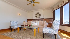 Una sala de estar con un sofá color beige, dos sillones estampados, una mesa de centro de vidrio, piso de madera, un ventilador de techo, un reloj en la pared y un amplio ventanal con vistas.