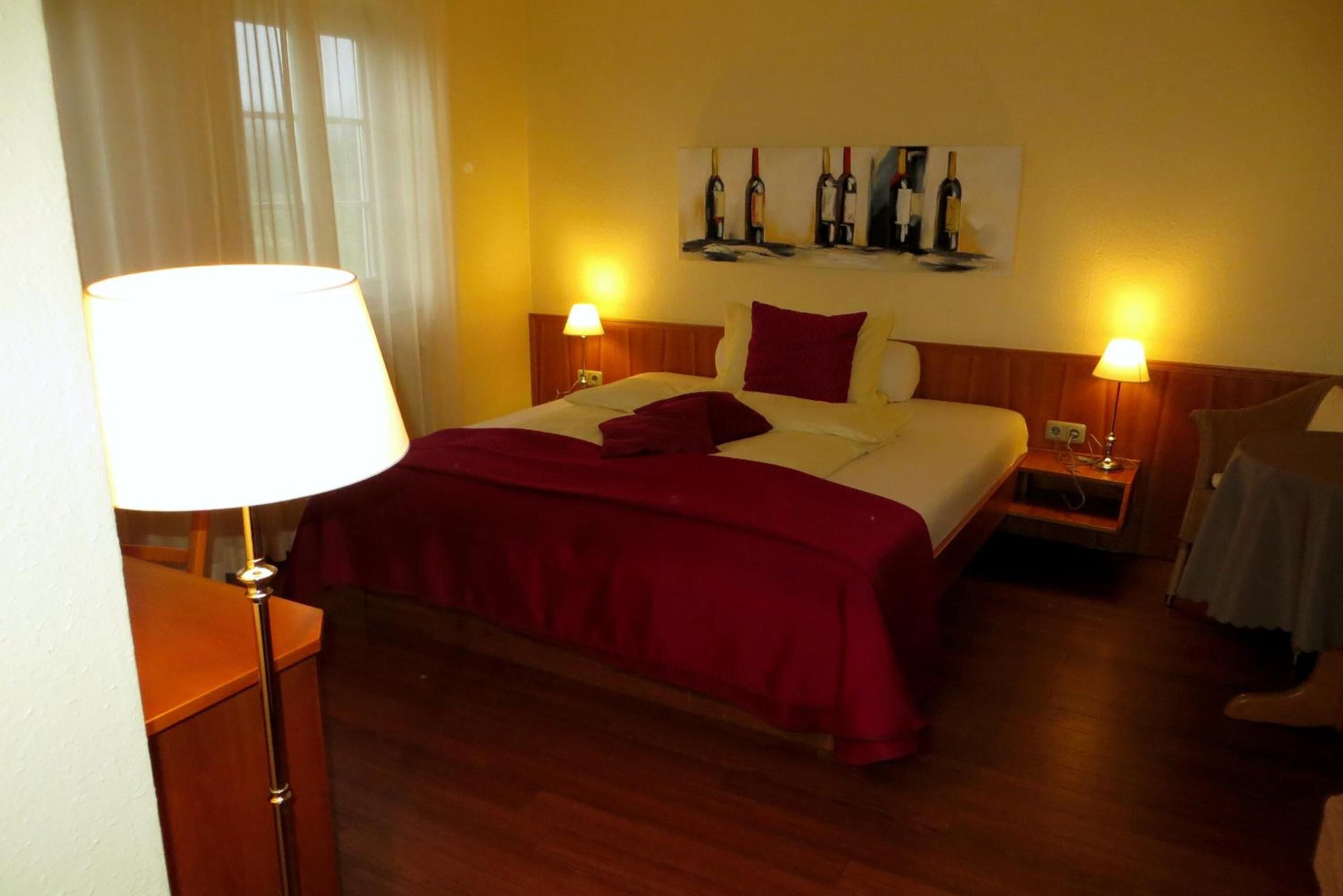 A hotel room with a bed, two bedside tables, a lamp, a painting of wine bottles, and a window with curtains.