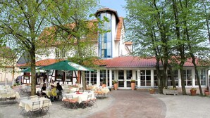 Un área de café al aire libre con mesas y sillas, un espacio de estar techado y un edificio con tejado de tejas rojas al fondo.