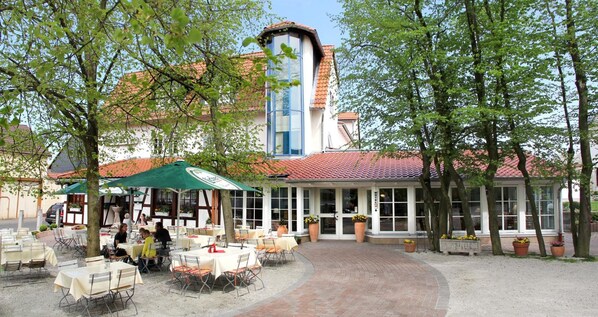 An outdoor cafe area with tables and chairs, a covered seating area, and a building with a red-tiled roof in the background.