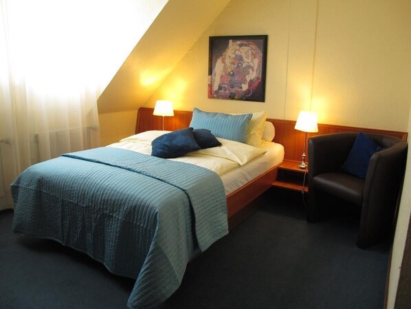 Habitación de hotel con cama, dos lámparas de noche, una silla y un cuadro en la pared.