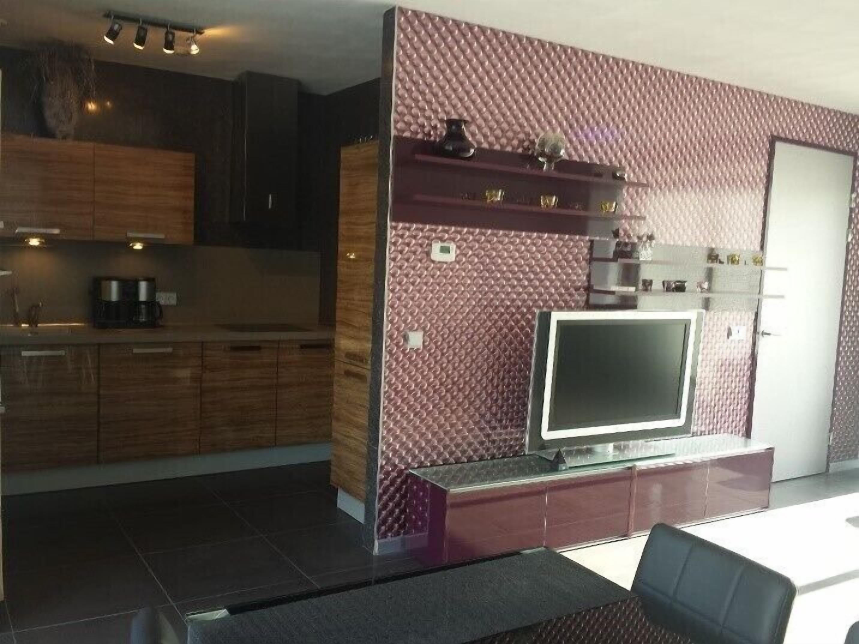 A modern kitchen with dark wood cabinets, a built-in oven, and a flat-screen TV mounted on a textured wall.