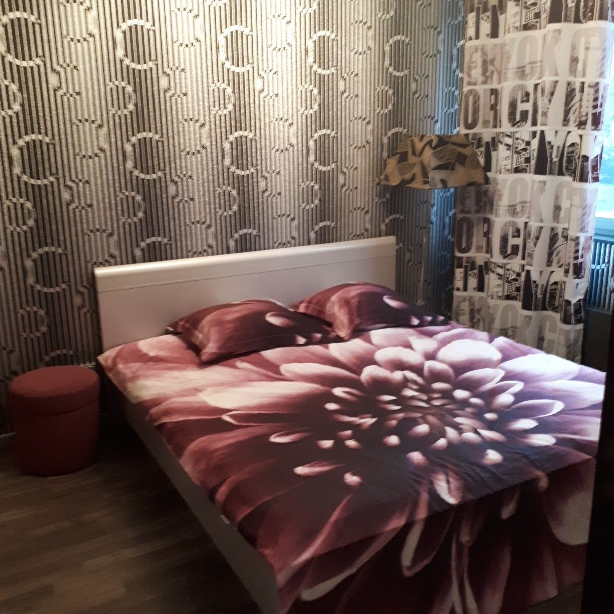 A bedroom with a bed, a red ottoman, patterned wallpaper, and a wall with printed material.