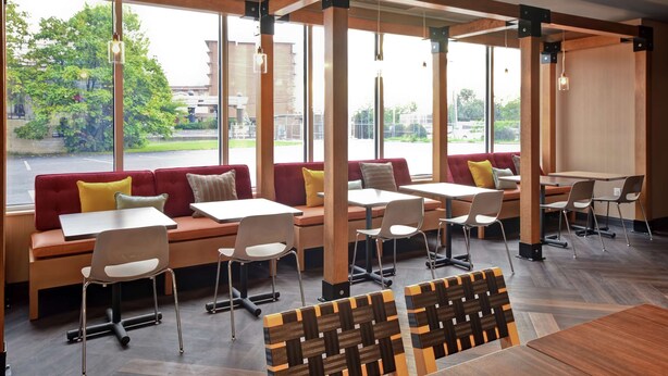 A modern indoor seating area with wooden tables and chairs, large windows offering a view of the outdoors, and a mix of red and beige seating. - Tru by Hilton Louisville Airport (Louisville)