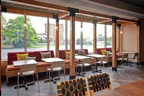 A modern indoor seating area with wooden tables and chairs, large windows offering a view of the outdoors, and a mix of red and beige seating. - Tru by Hilton Louisville Airport (Louisville)