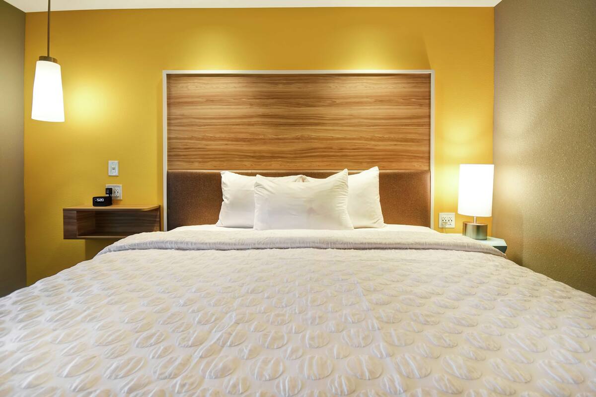 Une chambre d’hôtel avec un grand lit, une tête de lit en bois, une table de chevet avec une lampe et une étagère fixée au mur.