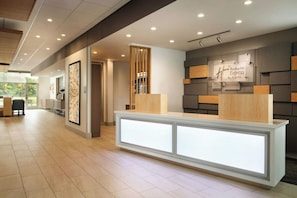 A hotel reception area with a modern design, featuring a reception desk, a wall-mounted sign for Holiday Inn Express & Suites, and a large window providing a view of the outdoors.