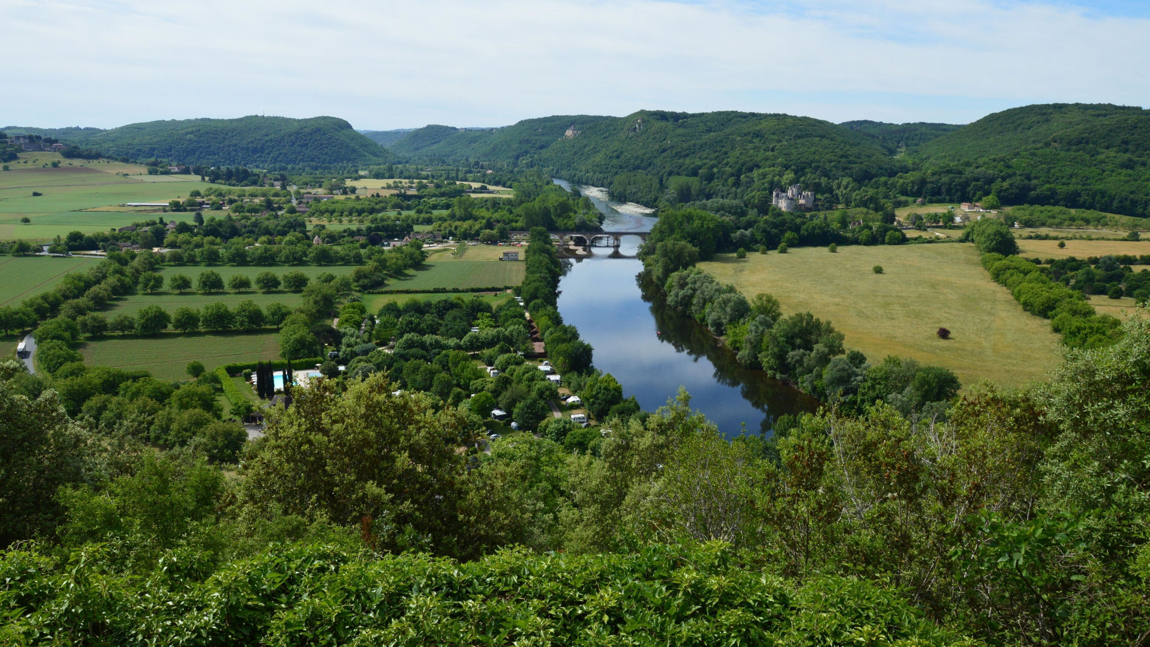 A river flowing through a valley with fields and trees, a bridge, and a castle-like structure in the distance.