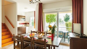 Uma sala de jantar com uma mesa de madeira posta para quatro, cozinha ao fundo, escada e varanda com vista para o exterior.