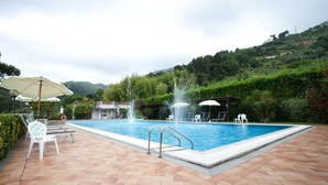 A swimming pool with a fountain, surrounded by lounge chairs and umbrellas, set against a backdrop of lush greenery and hills. - Hotel Ristorante Grappolo D'Oro (Pietrasanta)